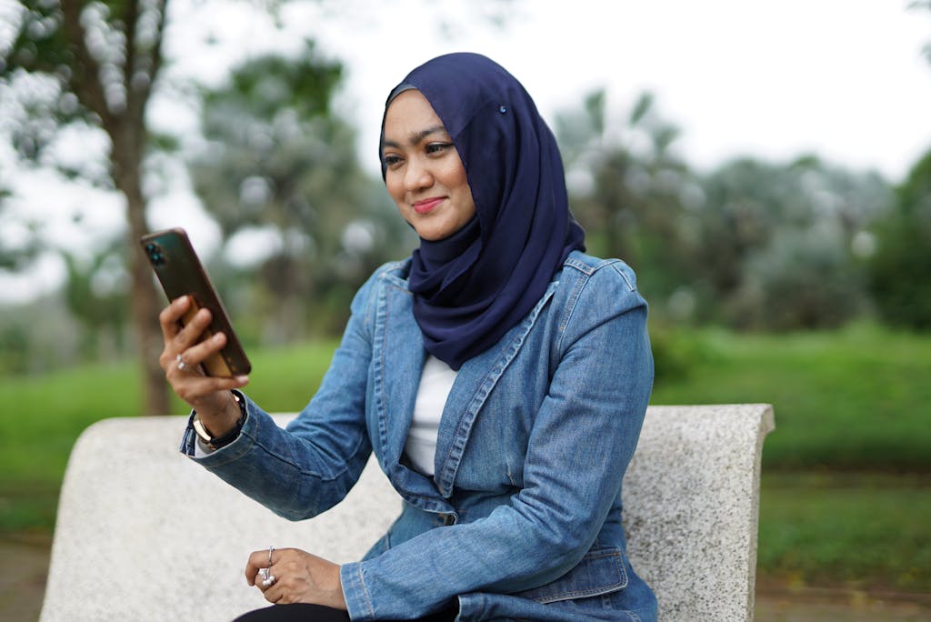 A cheerful woman in hijab using her phone while sitting on a bench in a park.