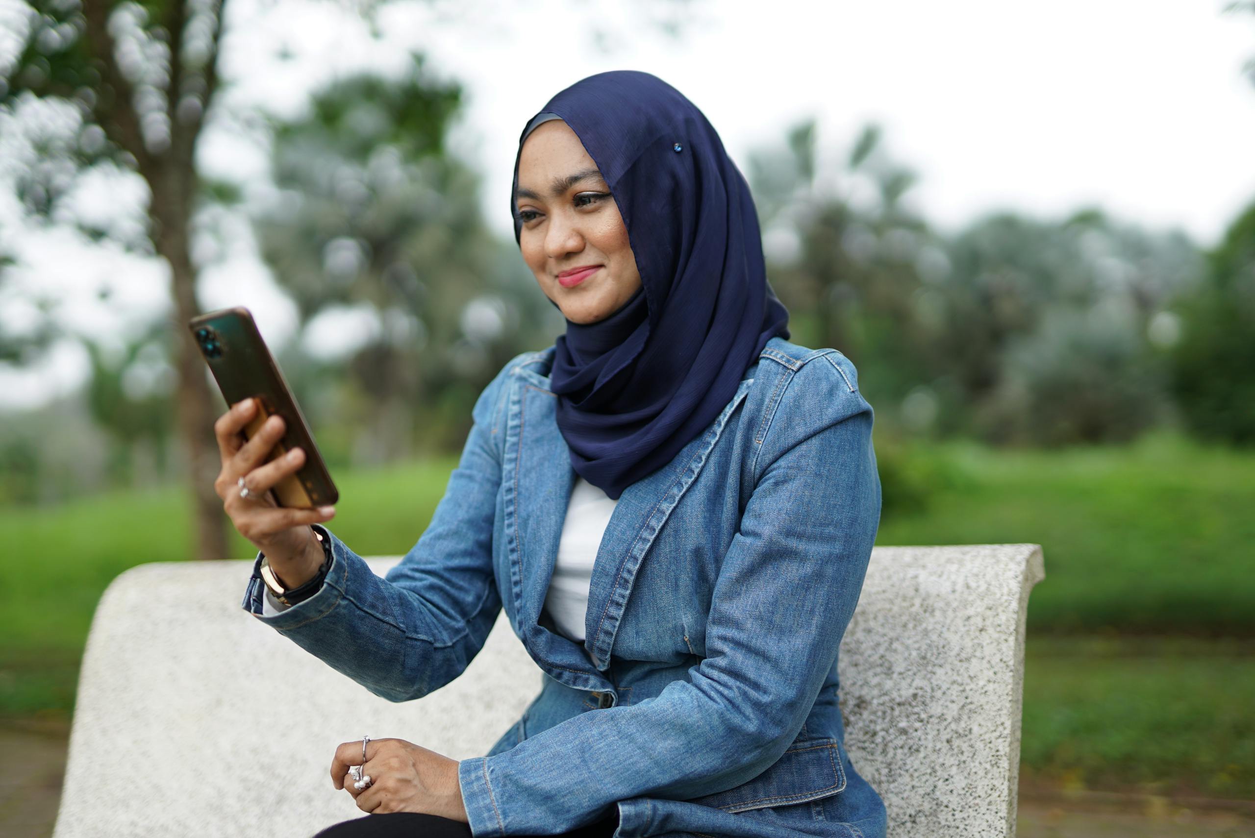 A cheerful woman in hijab using her phone while sitting on a bench in a park.
