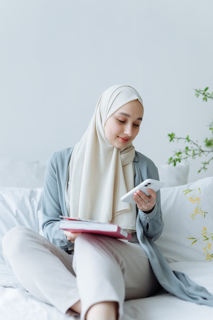 A young Muslim woman sits indoors, smiling as she reads on her phone. Calm and serene atmosphere.