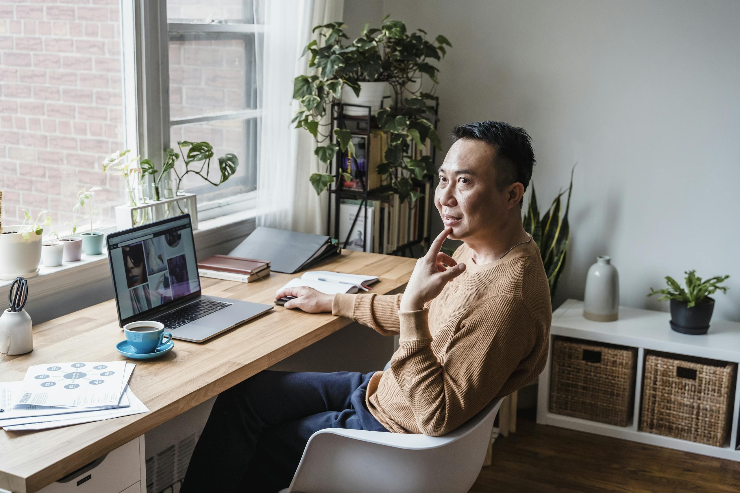 Asian man sitting at a desk with a laptop, coffee cup, and plants, working remotely indoors.