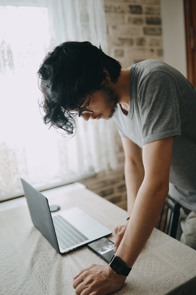 Focused young man leans over laptop working at home. Natural light filters through the window.