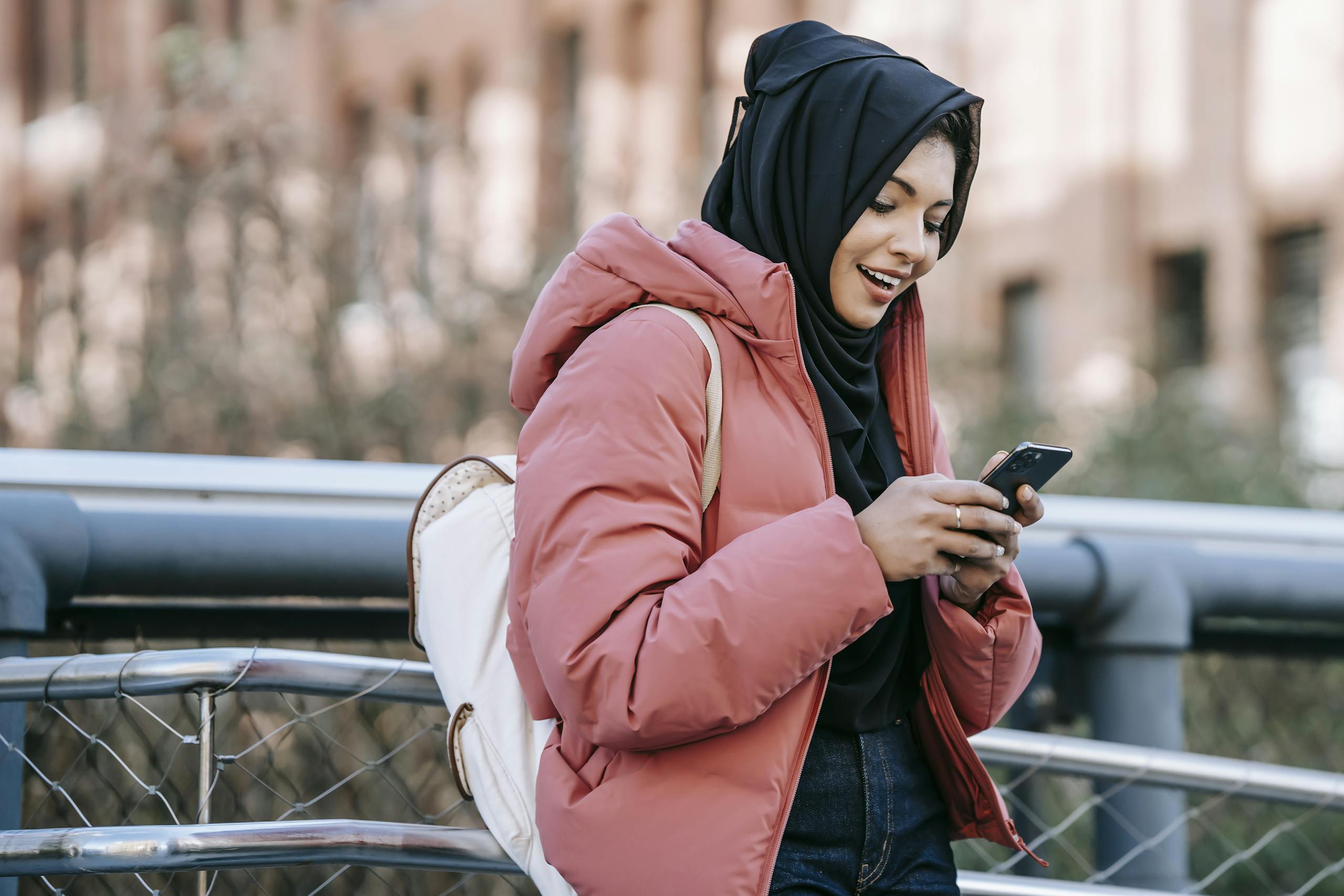 Muslim ethnic female in hijab texting message on mobile phone near iron construction on blurred background of building