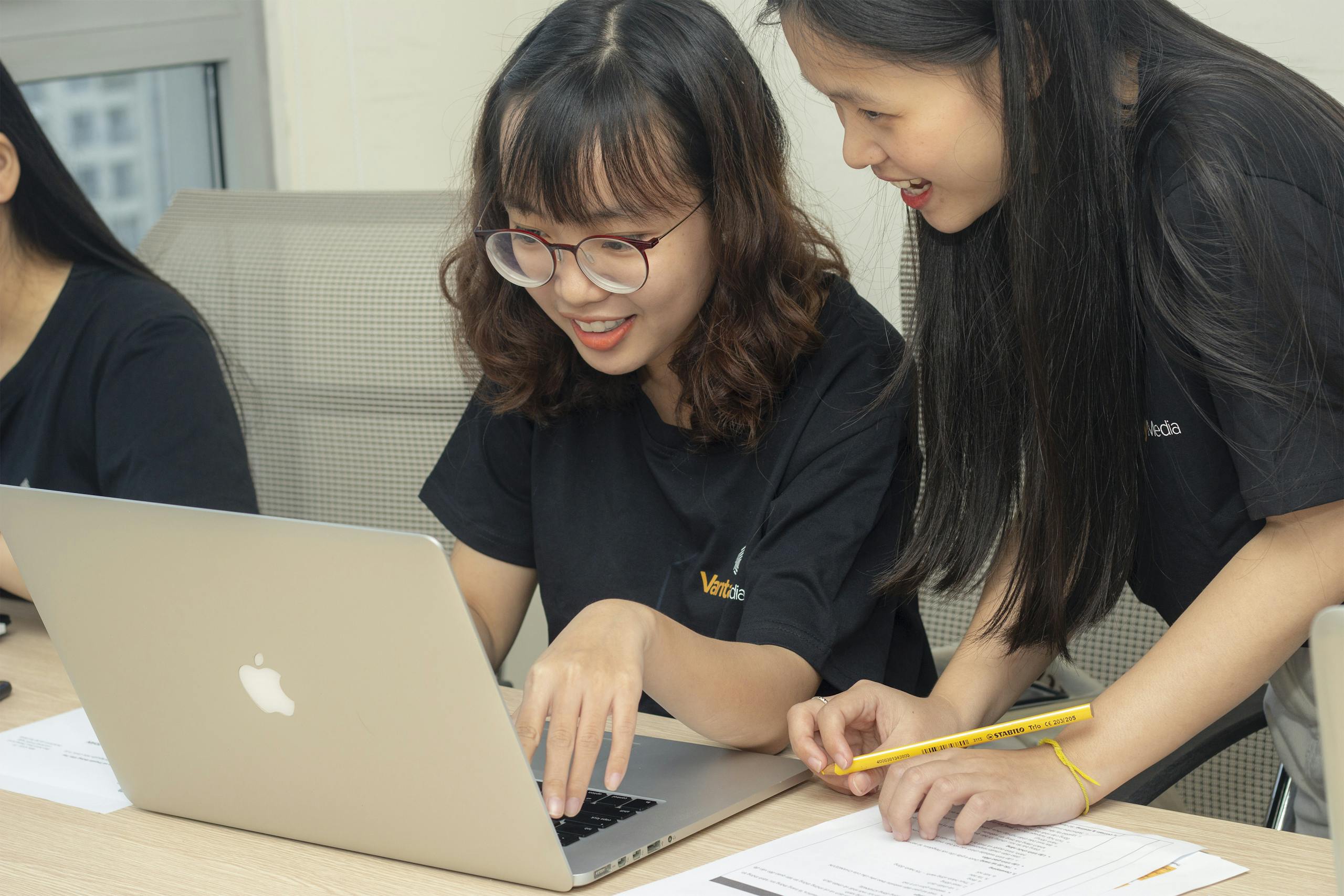 Two women smiling and working together on a laptop, embodying teamwork and collaboration in an office.